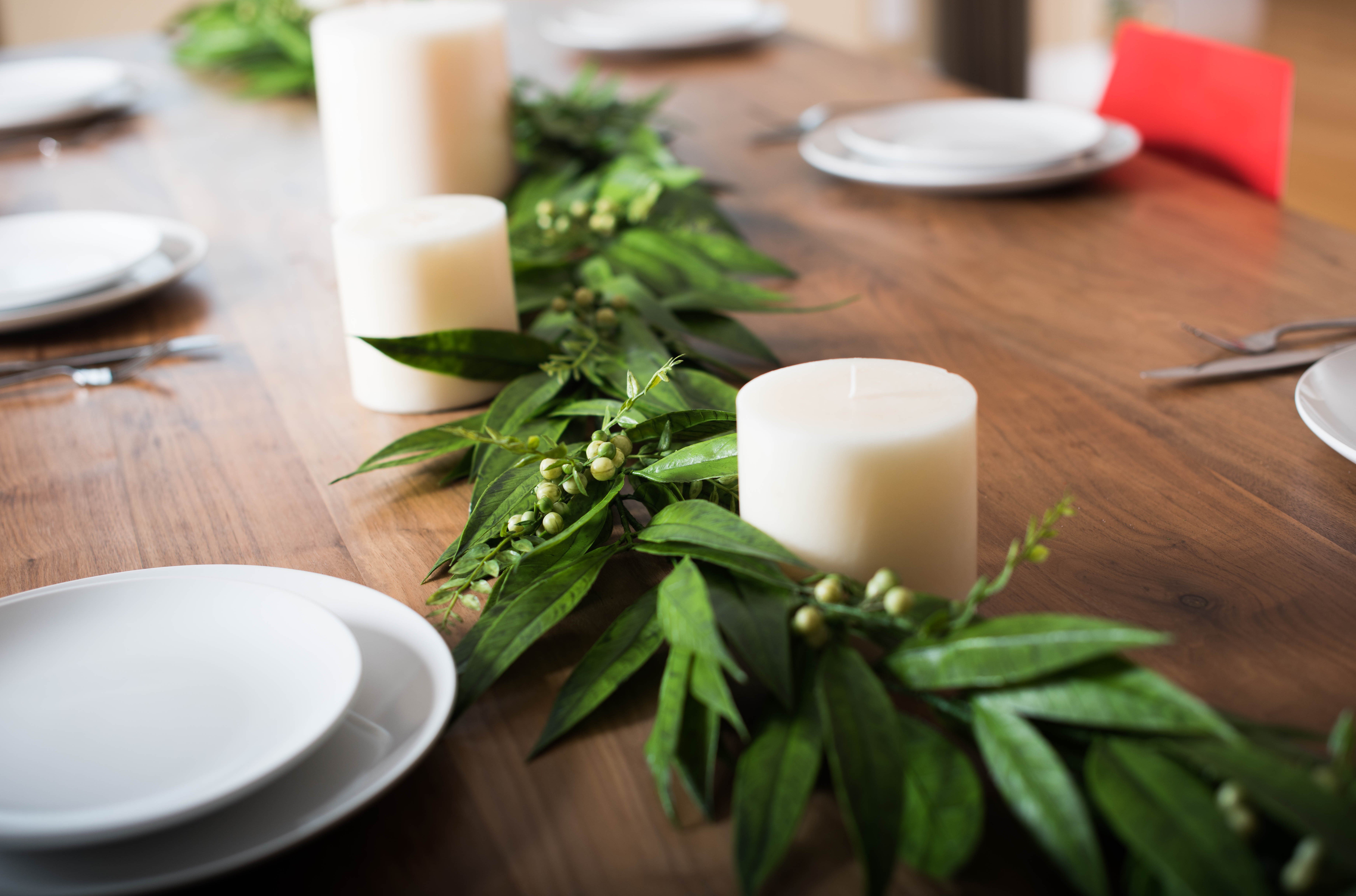 Cream unlit candles sit with green garland with green berries on wood table next to place settings